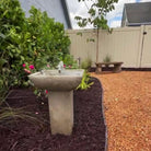 Andra cast stone fountain with bubbling water surrounded by flowering plants in a landscaped garden